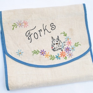 May include: A beige fabric pouch with blue trim featuring embroidered floral designs and a small dog illustration. The word "Forks" is stitched in black thread on the front flap. The pouch has a rounded flap closure and a textured, woven appearance.