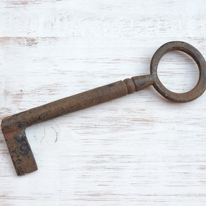 May include: An antique, rusted iron skeleton key featuring a large circular bow, a cylindrical shank with a decorative collar, and a simple L-shaped bit at the end. The metal surface shows significant oxidation, pitting, and a dark brown patina.