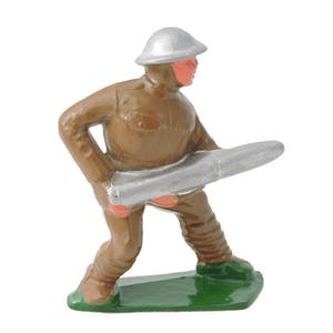 May include: A toy soldier figurine in a brown uniform and silver helmet, holding a silver object. The soldier stands on a green base, with red accents on the face and hands. The figurine is a vintage toy.