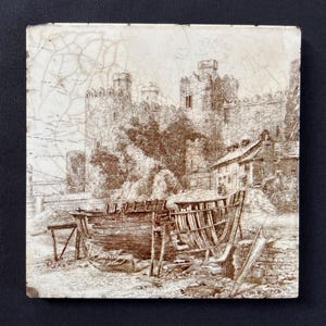 May include: A square ceramic tile featuring a sepia-toned line drawing of a stone castle with multiple towers and battlements. In the foreground, a wooden boat frame sits on a shore near a small cottage, all set against a white crackled glaze.