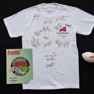 May include: White t-shirt with multiple signatures, the team logo, and the word "Piranhas" in red. A football and a program from the 1996 Inaugural Piranhas Season are also visible.