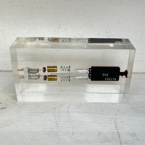 May include: A clear acrylic block encases disassembled mechanical components. The components include small motors, springs, and a black rectangular piece with the text "E02 12A1TA". The arrangement suggests an exploded view of a machine's inner workings.