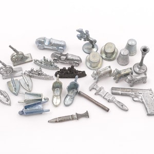 May include: A collection of silver-colored game pieces, including a cannon, race car, ship, top hat, thimble, and a pistol. The pieces are arranged on a white surface, showcasing a variety of shapes and sizes.