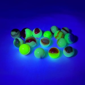 May include: A collection of glowing marbles in various colors, including green, yellow, red, and brown, against a blue background. The marbles have unique patterns and swirls, creating a visually interesting display. The image is lit with a black light.