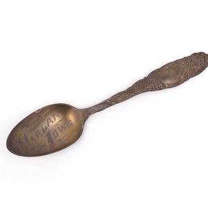 May include: A tarnished bronze-toned metal spoon featuring an ornate, embossed floral pattern along the handle. The bowl of the spoon displays the engraved text "HARLAN IOWA" in a dark, weathered font, showing signs of age and oxidation.