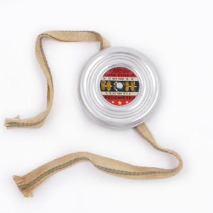 May include: A vintage silver pedometer with a beige fabric strap. The pedometer has a red and black face with the text "EMERSON COIL RANGE PEDOMETER". The strap is attached to the pedometer and is laying on a white surface.