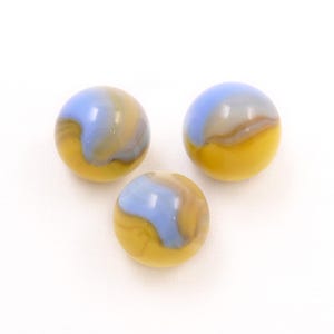 May include: Three glass marbles with a swirl design. Each marble features a combination of blue, yellow, and brown colors. The marbles are round and have a glossy finish, set against a white background.