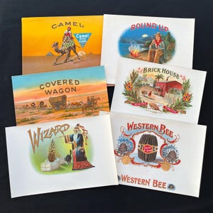 May include: A collection of vintage cigar box labels. The labels feature colorful illustrations and text, including images of a camel, a covered wagon, a wizard, and a cowboy. The labels are rectangular and vary in size.