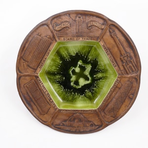 May include: A brown ceramic ashtray with a green, hexagonal center. The ashtray features embossed images of Chicago landmarks, including the Water Tower and John Hancock Center, and the text "CHICAGO ILLINOIS BOARD OF TRADE".