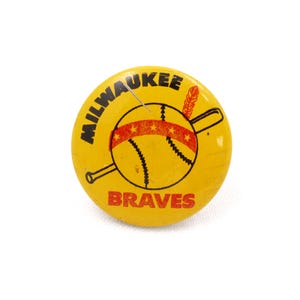May include: A yellow button with the words "MILWAUKEE BRAVES" in black lettering. The button features a baseball with a red and gold band, a feather, and a baseball bat. A vintage sports collectible.