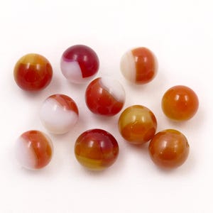 May include: A collection of ten glass marbles in shades of red, orange, and white. The marbles are round and have a glossy finish. Some have a swirl pattern, while others are solid in color. They are scattered on a white surface.