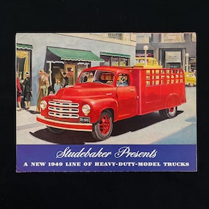 May include: A vintage advertisement for a red Studebaker truck. The truck has a distinctive grille and a cargo bed with a lattice design. The ad includes the text "Studebaker Presents" and "A New 1949 Line of Heavy-Duty-Model Trucks."