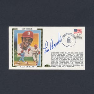 May include: A commemorative envelope featuring a portrait of baseball player Lou Brock, a blue ink autograph, and a 22-cent USA postage stamp postmarked Cooperstown, NY, July 28, 1985. The text details his induction into the Hall of Fame.
