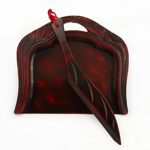 May include: A dark red lacquered dustpan and brush set featuring carved floral motifs along the top edge of the pan. The set includes a matching long, slender brush with carved leaf-like details and a small red cord loop for hanging the pan.