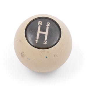 May include: A spherical beige gear shift knob featuring a circular black center insert. The insert displays a white H-pattern gear shift diagram with the letters R, 1, 2, and 3 clearly marked. The beige surface shows minor signs of wear and scuffing.