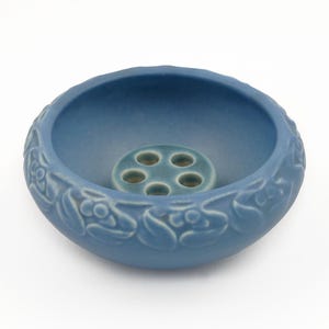 May include: A blue ceramic flower frog with a round shape and a central insert with six holes. The rim of the bowl is decorated with a repeating pattern of stylized frog faces. The frog is a decorative item.