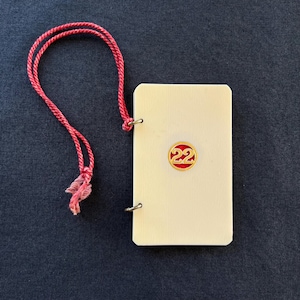 May include: Ivory rectangular tag with a red and gold emblem featuring the number 22. A red cord is attached to the tag with a loop at the top. The tag has a textured surface and is set against a dark blue background.