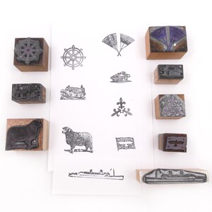 May include: A collection of wooden rubber stamps arranged around a central white paper sheet featuring black ink impressions of various designs including a ship's wheel, a sheep, a fleur-de-lis, a cruise ship, and other detailed graphic illustrations.