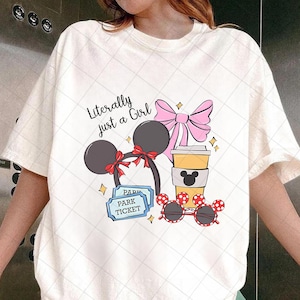 May include: Cream-colored t-shirt with a graphic design featuring a Minnie Mouse ear headband, a pink bow, a coffee cup, sunglasses, and park tickets. The text "Literally just a Girl" is also included.