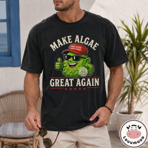 May include: A black short-sleeve t-shirt featuring a graphic of a green, textured algae character wearing a red cap and black sunglasses. The text reads "MAKE ALGAE GREAT AGAIN" with five red stars below the text. The character gives a thumbs up.