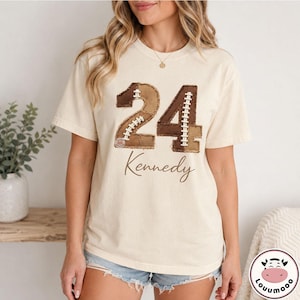 May include: A cream-colored short-sleeve t-shirt featuring a large graphic of the number 24 designed to look like a football with brown textured panels and white laces. The name Kennedy is printed in a brown cursive script below the number.