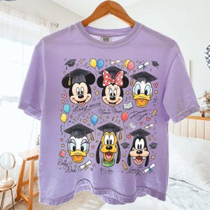 May include: A lilac-colored t-shirt featuring Mickey Mouse, Minnie Mouse, Donald Duck, Daisy Duck, Pluto, and Goofy, all wearing graduation caps. The design includes balloons, books, and diplomas.