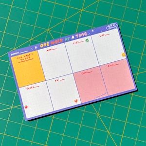 Pode incluir: Um planejador semanal com uma borda roxa e um fundo azul claro. O planejador é dividido em sete seções para cada dia da semana, com uma seção amarela para "O que fazer esta semana". O planejador tem um padrão de pontos e inclui pequenas ilustrações de uma flor, um morango, um coração e um rosto sorridente.