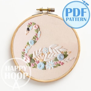 May include: An embroidered swan design within a wooden embroidery hoop. The swan is adorned with pink, blue, and white floral details and green leaves, set against a pale pink backdrop. The text "PDF PATTERN" is displayed.