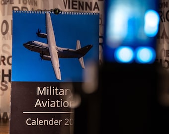 May include: A wall calendar featuring a photograph of a gray military transport aircraft in flight against a blue sky. The lower portion is black with the white text Military Aviation Calender 20. The calendar is spiral bound at the top.