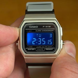 Puede incluir: Reloj digital Casio F-91W plateado con correa gris. La esfera del reloj muestra la hora 2:35:15. El reloj tiene el texto "CASIO F-91W" y "WATER RESIST" impreso en la esfera.