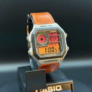 May include: A Casio digital wristwatch with a silver-tone square case and a textured orange leather strap. The watch face features a world map display, multiple alarm indicators, and an orange digital time readout labeled World Time and Illuminator.