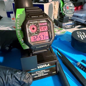 May include: A black Casio digital watch with a rectangular face displaying the time in pink digits. The watch has multiple functions, including world time and alarms. A green tag reads "10 Year Battery". Another Casio watch is visible in the background.