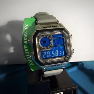 May include: A digital Casio watch with a silver-tone case and a blue digital display showing the time. The watch has a gray band and a green tag that reads "10 YEAR BATTERY". The watch is displayed on a black stand.
