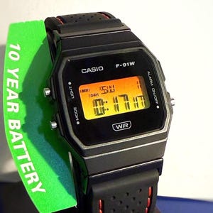 Op de afbeelding: Zwarte Casio F-91W digitale horloge met een rechthoekige wijzerplaat en een zwarte band met rode accenten. Het horloge toont de tijd op een oranje digitaal scherm. Een groen label toont "10 YEAR BATTERY".