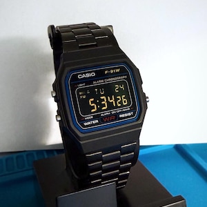 Può includere: Orologio digitale Casio F-91W nero con quadrante rettangolare e cinturino in metallo nero. Il quadrante mostra l'ora, il giorno e la data in formato digitale. L'orologio è su un espositore nero. L'orologio ha il testo "CASIO" e "F-91W" sul quadrante.