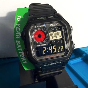 May include: Black Casio digital watch with a rectangular face. The watch displays the time, date, and world time features. The watch has a red dial and a green tag that reads "10 YEAR BATTERY".