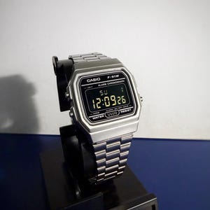 Könnte beinhalten: Eine silberne Casio F-91W Digitaluhr mit passendem Metallarmband. Die Uhr zeigt die Zeit 12:09:26 auf einem schwarzen Bildschirm an. Die Uhr steht auf einem schwarzen Ständer. Das Casio-Logo ist sichtbar.