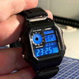 May include: Black Casio digital watch with a square face and a black band. The watch face displays the time, date, and other functions in blue. The watch has buttons labeled "LIGHT" and "SEARCH". The watch is water resistant to 100 meters.