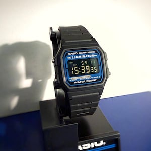 May include: Black Casio digital watch with a blue and black display. The watch shows the time 15:39:35. The watch has the text "CASIO ALARM CHRONO ILLUMINATOR WATER RESIST" on the face. The watch is on a black display stand.