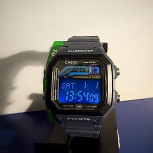 May include: A digital Casio watch with a blue display showing the date and time. The watch has a dark gray case and band. Features include an alarm, countdown timer, and 10-year battery. The watch is displayed on a black surface.