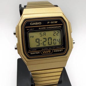 May include: Gold-toned Casio F-91W digital watch. The watch face displays the time as 9:20:04 PM on Saturday, the 22nd. Features include alarm, chronograph, and water resistance. The watch is displayed on a black stand.