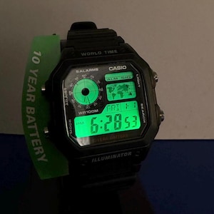 May include: Black digital watch with a green illuminated display showing the time and world map. The watch has multiple buttons and features, including world time and alarm functions. A green band with "10 YEAR BATTERY" is visible.