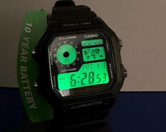 Puede incluir: Un reloj digital Casio negro con una pantalla retroiluminada de color verde brillante que muestra la hora, la fecha y un mapa mundial. El reloj cuenta con una subesfera analógica redonda, una pantalla digital rectangular y una etiqueta verde 10 YEAR BATTERY.