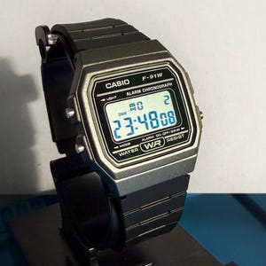 Op de afbeelding: Een zwarte Casio F-91W digitale horloge met een zilverkleurige rand. Het horloge toont de tijd, datum en andere functies op een digitaal scherm. Het heeft een zwarte harsband en is waterbestendig.