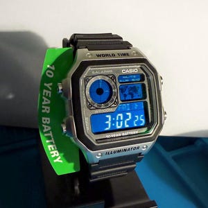 May include: A Casio digital watch with a black strap and silver-tone case. The watch face displays the time in blue digits and includes the text "WORLD TIME" and "10 YEAR BATTERY". A green tag with "10 YEAR BATTERY" is attached.