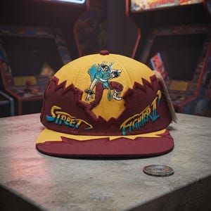 Puede incluir: Una gorra de béisbol amarilla y granate con un gráfico bordado de un personaje de Street Fighter II. La gorra muestra el texto Street Fighter II en bordado azul y amarillo en la parte frontal, con un patrón de diseño en zigzag.
