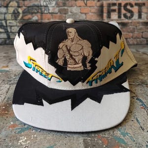 Puede incluir: Una gorra de béisbol negra y color crema con una figura musculosa bordada en el panel frontal. La gorra muestra el texto Street Fighter en bordado azul y amarillo. El diseño incluye patrones de bloques de colores contrastantes.