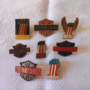 Może przedstawiać: Kolekcja przypinek Harley-Davidson w różnych kształtach. Przypinki przedstawiają logo Harley-Davidson, numer jeden i wzory flagi amerykańskiej. Kolory obejmują czarny, czerwony, pomarańczowy, biały i niebieski. Przypinki są wyświetlane na białej, teksturowanej powierzchni.