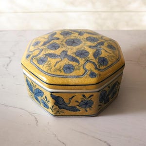 Könnte beinhalten: Gelbe, sechseckige Keramik-Schmuckschatulle mit Deckel. Verziert mit blauen Blumen- und Vogelmotiven. Mit weißem Rand und Craquelé-Finish.