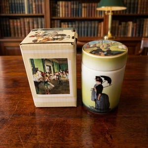 Puede incluir: Un tarro cilíndrico de porcelana con tapa, con un acabado en degradado verde y blanco. La tapa muestra una escena colorida al aire libre, mientras que el lateral representa una figura con vestido negro sosteniendo un paraguas negro. Se encuentra junto a su caja.